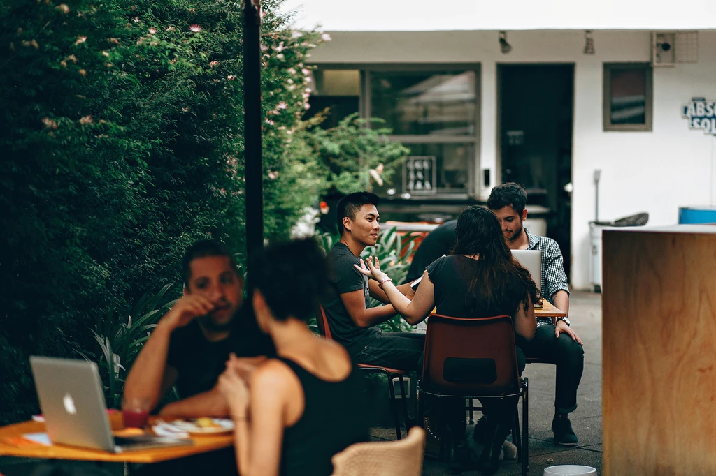Pessoas trabalhando e reunidas em mesas ao ar livre de um café, algumas com laptops